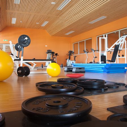 Ein Fitnessstudio mit Holzböden, orangefarbenen Wänden und verschiedenen Geräten wie Langhanteln, Kurzhanteln und Übungsbällen. Das Fitnessstudio verfügt über verschiedene Trainingsgeräte und blaue Matten auf dem Boden.