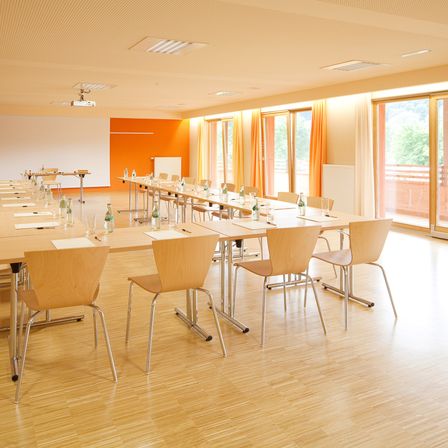 Ein Konferenzraum mit Holzboden, langen Tischen, Stühlen und großen Glasfenstern. Es hat einen Projektor und eine Tafel. Der Raum ist leer.
