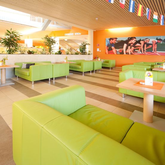 Ein heller, moderner Empfangsbereich mit lindgrünen Sofas um Tische herum, betont durch orange Wände und ein großes Wandbild. Dekorative Flaggen hängen von der Decke.
