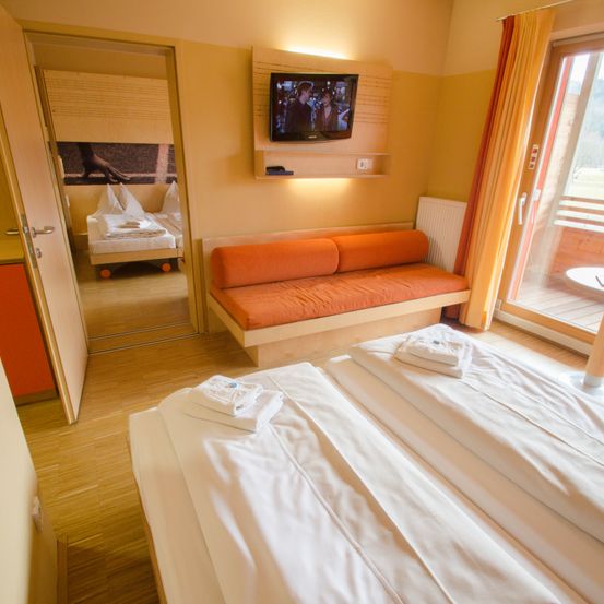 Ein Schlafzimmer mit einem Doppelbett, einer Couch, einem Fernseher und einem Spiegel, der ein anderes Bett reflektiert. Der Raum hat Holzböden, und draußen ist eine Terrasse.