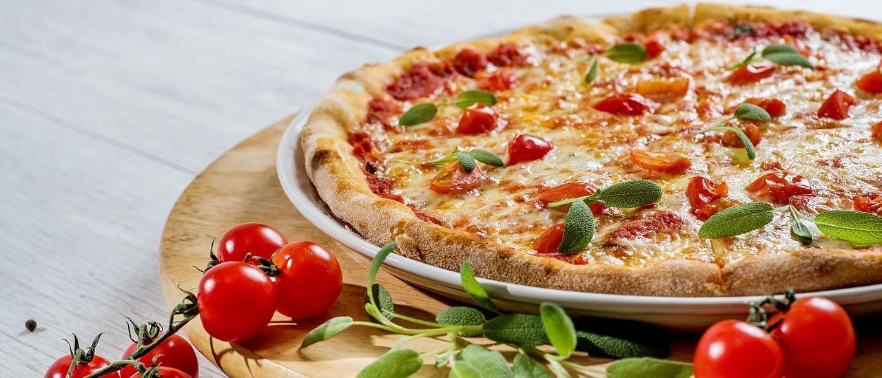 Eine ganze Pizza mit Käse, Tomaten und Salbeiblättern auf einem weißen Teller liegt auf einem Holztablett mit Kirschtomaten und Pfeffer auf einem Holztisch.