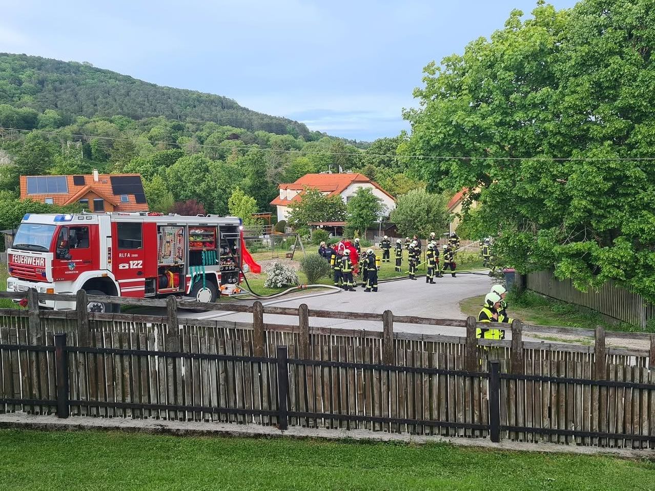 Ein roter Feuerwehrwagen ist am Straßenrand geparkt, mit Feuerwehrleuten um ihn herum. Sie tragen Schutzkleidung und Helme. Eine Person steht in der Nähe des Zauns, und ein Mülleimer ist in der Nähe. Das Gebiet ist von Bäumen und Häusern umgeben.