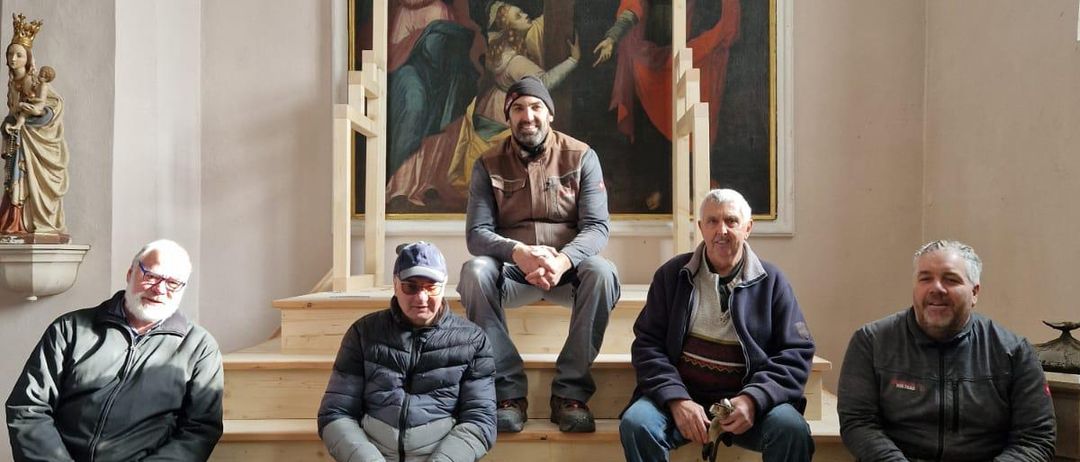 Fünf Männer sitzen auf den Stufen in einer Kirche. Sie lächeln und posieren für ein Foto. Ein Gemälde der Kreuzigung hängt an der Wand hinter ihnen.