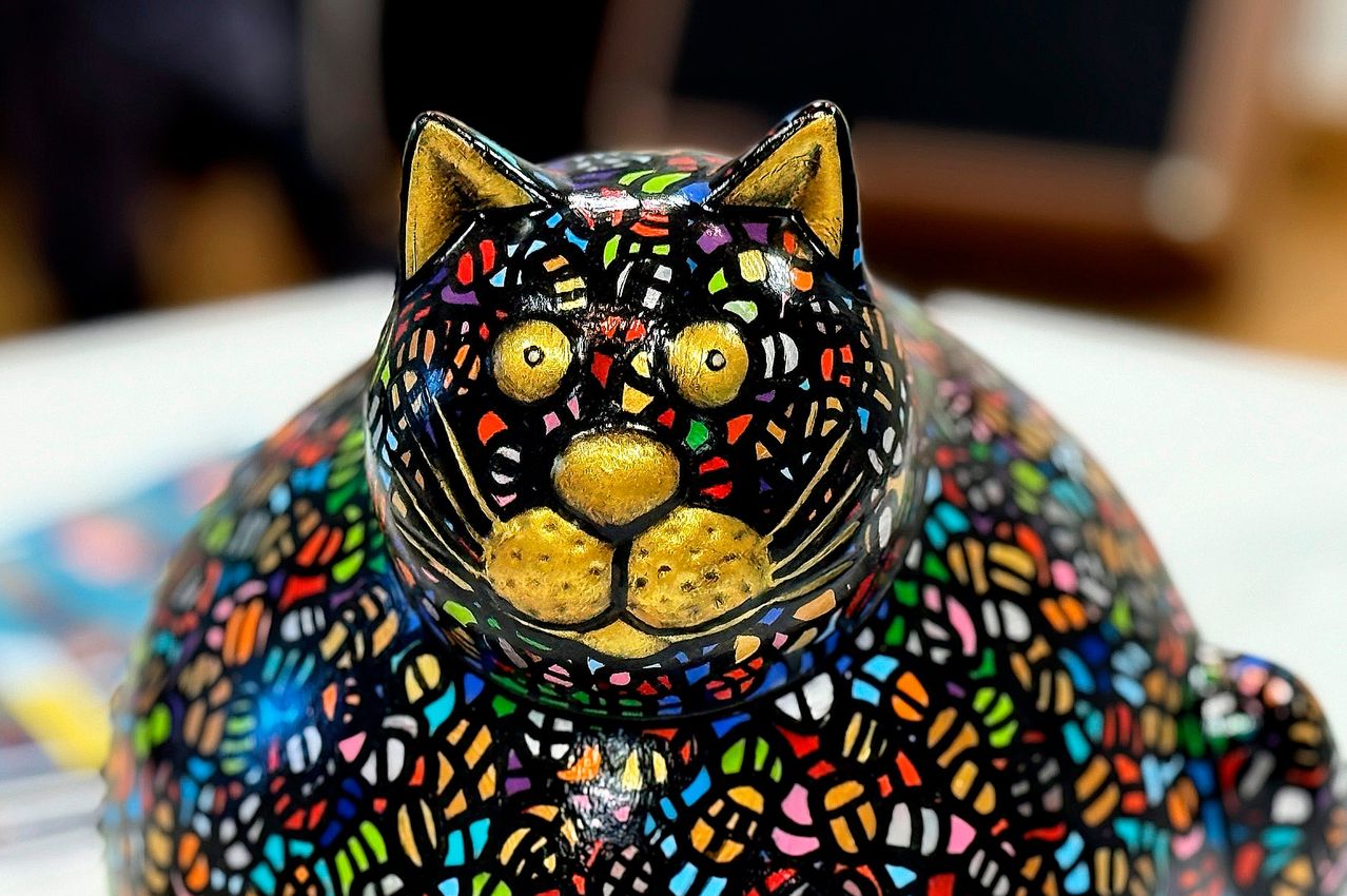 A close-up of a colorful ceramic sculpture of a cat with golden accents and a mosaic pattern. The cat's face is detailed with yellow eyes and a golden nose.