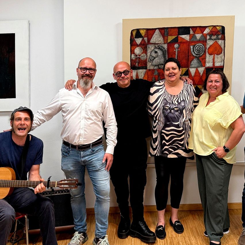 A group of people standing in an art gallery with a guitar player sitting on a chair. The man in the middle is wearing glasses and a necklace.