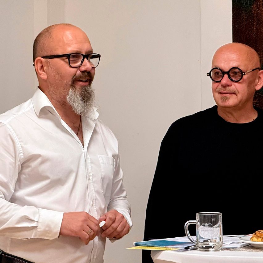 Two men, one with a beard and glasses, smiles. The other man wears a black shirt. They sit at a table with food and drink.