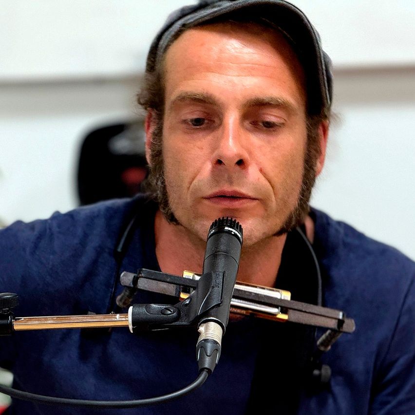 A man with a beard plays a harmonica into a microphone while wearing a hat and a blue shirt.