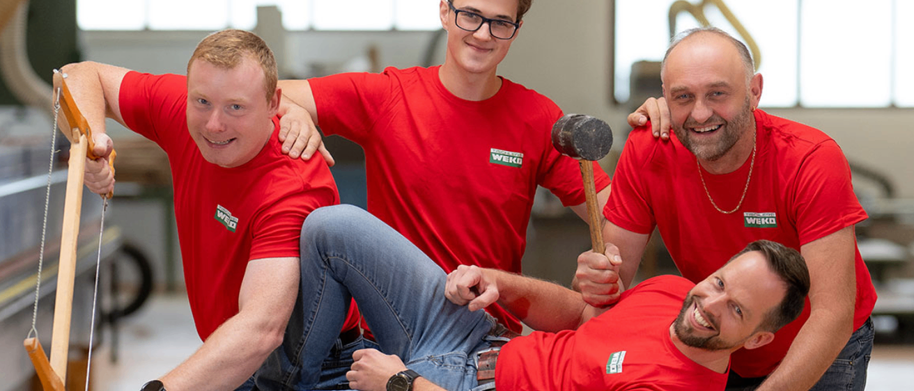 Vier Männer in roten T-Shirts, zwei stehend und zwei sitzend, lächeln. Einer hält einen Hammer. Drinnen, Tageslicht.
