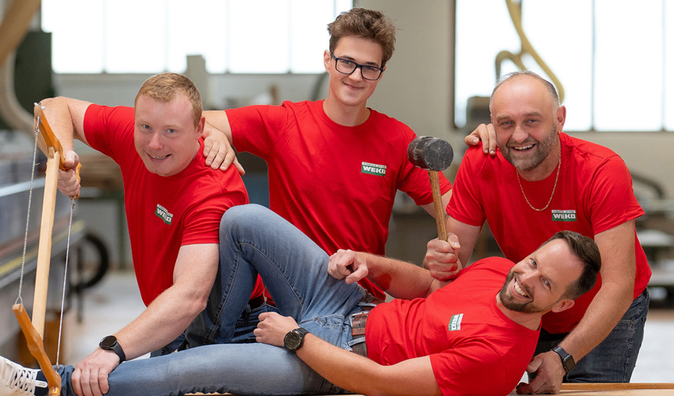 Vier Männer in roten T-Shirts, einer hält einen Hammer, posieren für ein Foto in einer Werkstatt.