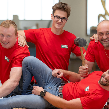 Vier Männer in roten T-Shirts, einer hält einen Hammer, posieren für ein Foto in einer Werkstatt.