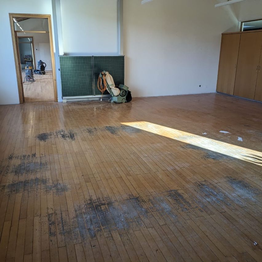 Ein leerer Raum mit Holzböden, Wänden und einem grünen Gitter. Reinigungsgeräte sind auf dem Boden platziert, und Sonnenlicht erhellt den Raum.