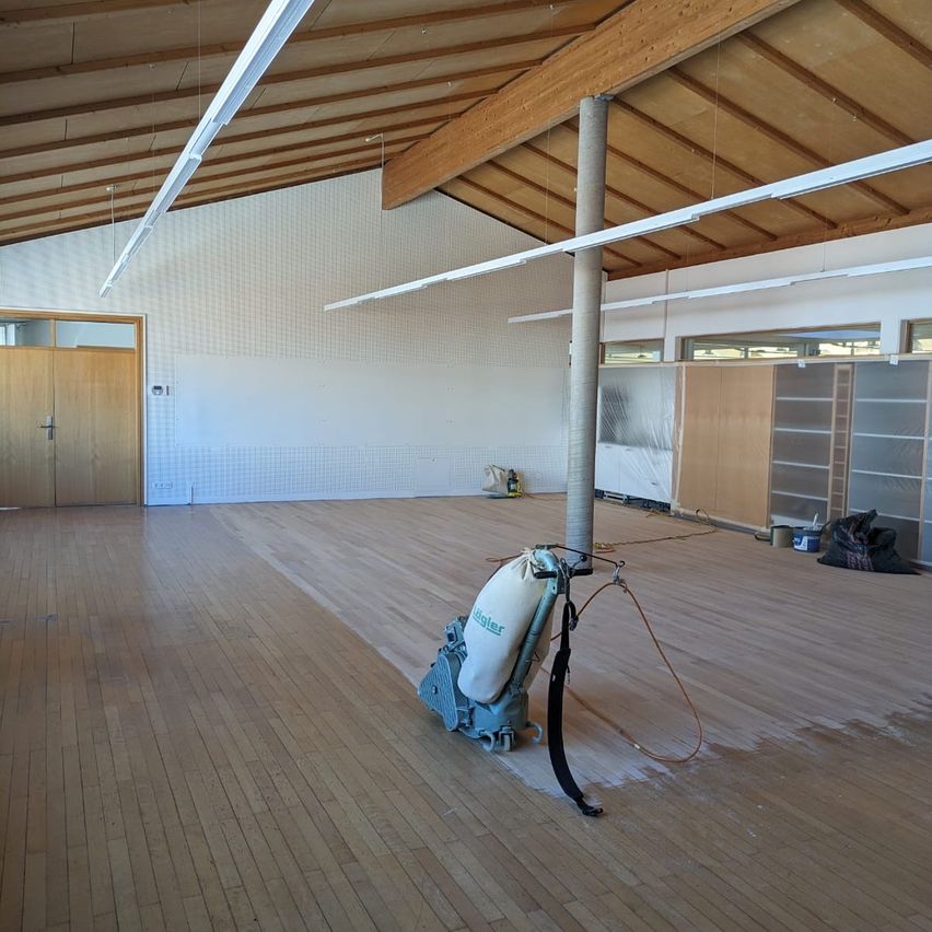 Ein leerer Raum mit Holzböden wird von einer großen Maschine geschliffen. Der Raum hat weiße Wände, Holzbalken und Leuchtstofflampen. Es gibt Regale und eine Tür auf der linken Seite.