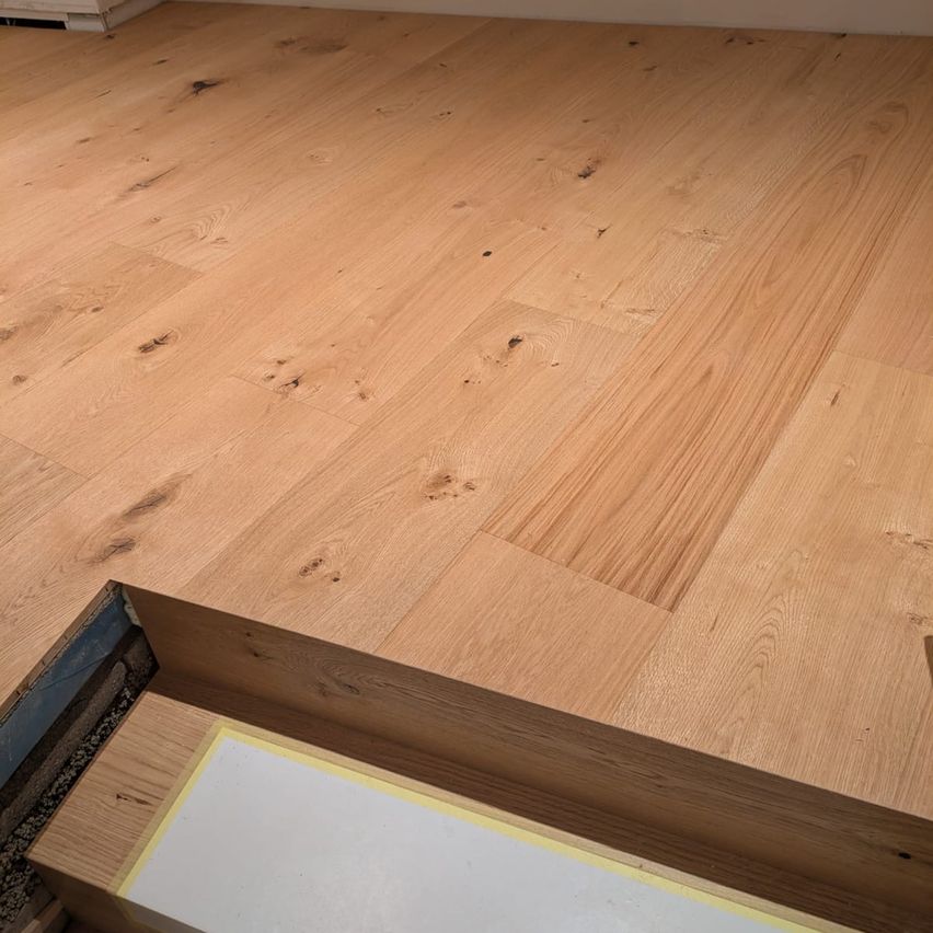 Eine Nahaufnahme eines Holzbodens mit sichtbaren Maserungen und einigen Unvollkommenheiten. Der Boden scheint unbehandelt zu sein und zeigt seine natürliche Textur.