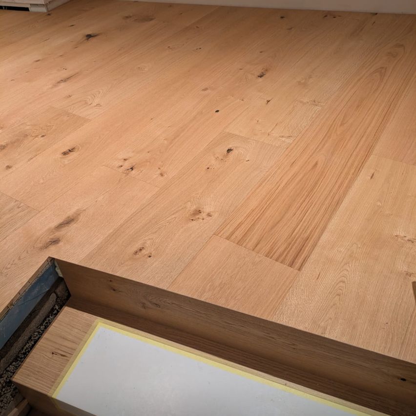 Eine Nahaufnahme eines Holzbodens mit sichtbaren Holzmaserungen und Knoten. Der Boden hat eine natürliche, hellbraune Farbe. Es gibt leichte Verfärbungen und kleine Löcher, die über die Oberfläche verstreut sind.