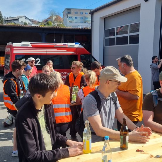 Eine Gruppe von Menschen in orangefarbenen Westen versammelt sich um einen Tisch mit Flaschen und Dosen. Ein Mann mit Hut sitzt am Tisch, während andere hinter ihm stehen. Ein Feuerwehrwagen ist hinter der Gruppe geparkt.