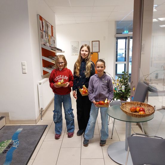 Drei Mädchen stehen in einem Flur und halten Körbe mit Obst. Sie lächeln und posieren für ein Foto.