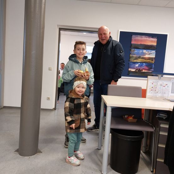 Ein Mann steht mit zwei Kindern, die Essen halten. Sie sind in einem Raum mit einem Schreibtisch, einem Stuhl und einem Mülleimer. Eine weitere Person steht in der Nähe einer offenen Tür.
