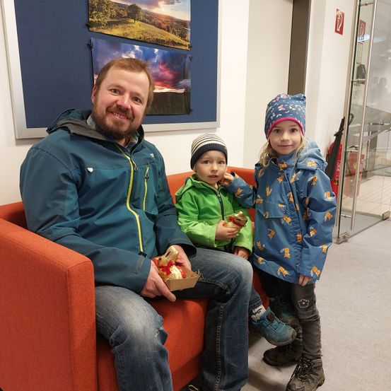 Ein Mann und zwei Kinder sitzen auf einem roten Sofa und halten Schokolade. Das Kind auf der linken Seite trägt eine Mütze und das Kind auf der rechten Seite trägt einen Hut. Sie alle lächeln. Dahinter befindet sich eine Wand mit gerahmten Bildern.