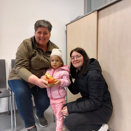 Eine Frau und ein Mädchen halten einen Korb mit Äpfeln und einem Schokoladenei. Eine weitere Mädchen mit Brille kniet neben ihnen auf dem Boden.