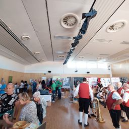 Ein Saal voller Menschen, viele von ihnen ältere Personen, die sich an verschiedenen Aktivitäten beteiligen. Einige stehen, andere sitzen. Eine Band spielt Musik, und es gibt mehrere Tische mit Gegenständen darauf. Die Decke hat Lüfter und Lichter.