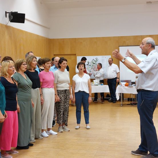 Eine Gruppe von Menschen, möglicherweise beim Singen, steht in einem Raum mit Holzböden und Wänden. Ein Mann führt sie an und gestikuliert mit den Händen. Ein Tisch mit Gegenständen steht in der Nähe.