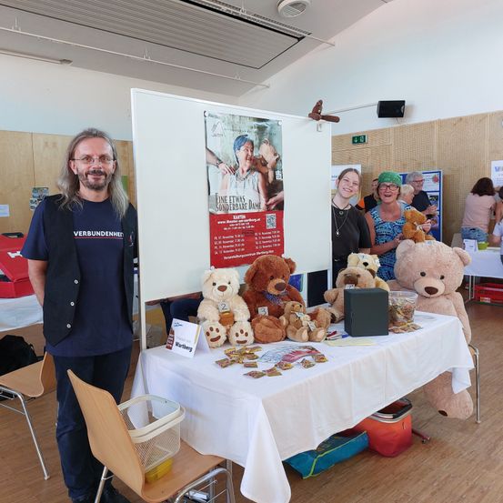 Ein Mann steht vor einem Stand mit Kuscheltieren, einem Plakat und einer weißen Tischdecke. Zwei Frauen stehen lächelnd hinter dem Tisch.
