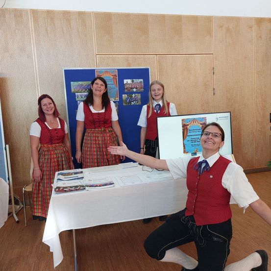 Drei Frauen in traditionellen Kostümen stehen neben einem Tisch mit Broschüren und lächeln, während ein Mann in einem roten Weste und Brille auf eine Tafel zeigt.