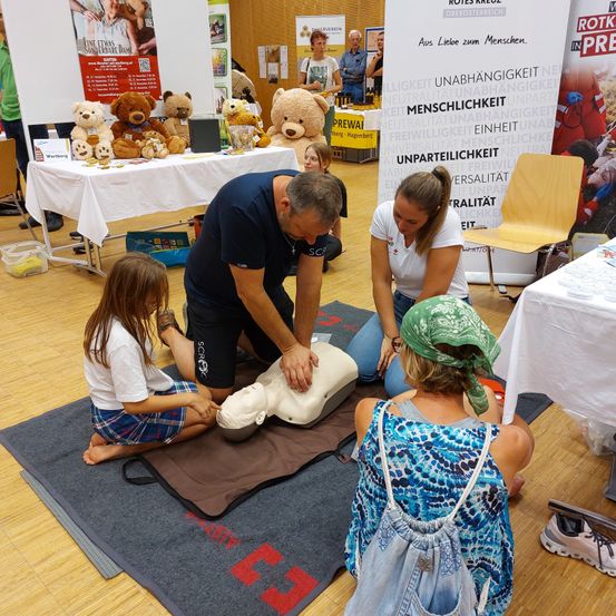 Ein Mann unterrichtet zwei Mädchen in der Durchführung von CPR an einem Dummy, während eine weitere Frau zusieht. Dahinter befinden sich Tische mit Kuscheltieren und Postern.