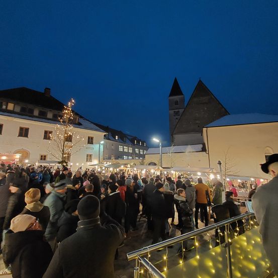 Ein Nachtmarkt ist bei einem dunklen Himmel voller Menschen vor Gebäuden mit Weihnachtsbeleuchtung, in der Nähe einer Kirche.