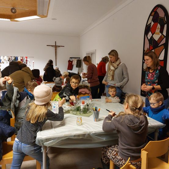 Mehrere Kinder und Erwachsene sind um einen Tisch in einem Raum versammelt. Einige malen oder zeichnen. Eine erwachsene Frau unterrichtet.