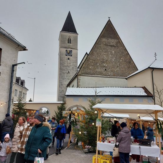 Ein Weihnachtsmarkt ist voller Menschen vor einer Kirche, geschmückt mit Schnee und Weihnachtsbäumen. Straßenlaternen und ein Glockenturm sind sichtbar.