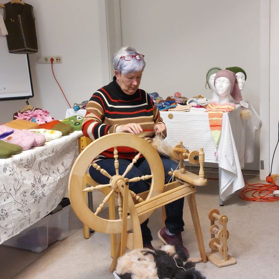 Eine ältere Frau mit Brille und gestreifter Strickjacke sitzt an einem Spinnrad und spinnt Garn. In der Nähe steht ein Tisch mit weißer Tischdecke, auf dem verschiedene Gegenstände wie Hüte ausgestellt sind. Im Hintergrund sind ein Whiteboard und Steckdosen zu sehen.