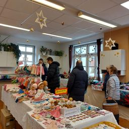 Eine Marktszene mit mehreren Personen, die Babykleidung und Spielzeug betrachten. Die Verkaufstische sind mit weißem Stoff bedeckt, und dekorative Sterne und Lichter schmücken die Decke.