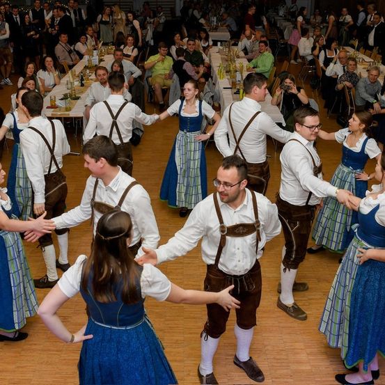 Mehrere Menschen tanzen in einem Saal, gekleidet in traditioneller deutscher Kleidung. Die Gruppe besteht aus Männern und Frauen, einige halten sich an den Händen. Der Raum ist mit anderen Menschen gefüllt, die an Tischen sitzen.