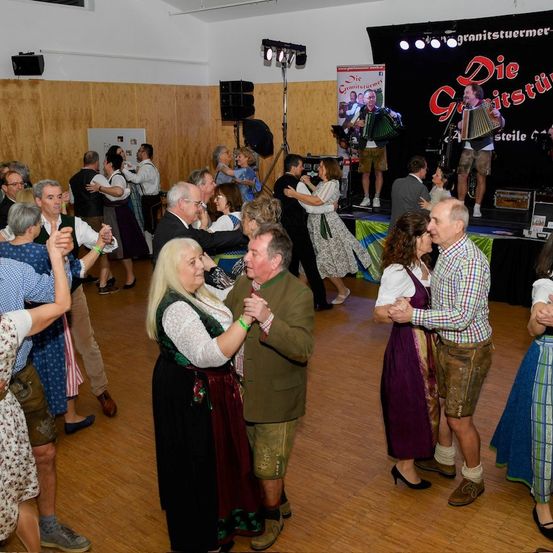 Eine Gruppe von Menschen tanzt in einem Saal. Zwei Personen spielen Instrumente auf der Bühne. Der Mann spielt Akkordeon und die Frau singt. Der Saal hat Holzboden und Holzwände. Es gibt Lautsprecher und ein Banner an der Wand.