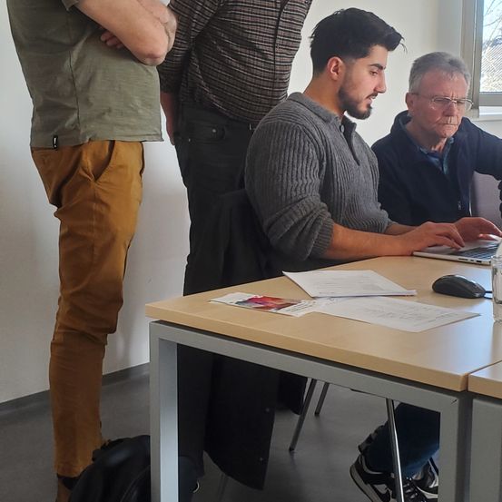 Drei Männer befinden sich in einem Raum mit einem Schreibtisch. Der Mann in der Mitte sitzt und tippt auf einem Laptop. Der Mann rechts trägt eine Brille und schaut den Mann in der Mitte an. Der Mann links steht und schaut den Mann in der Mitte an.