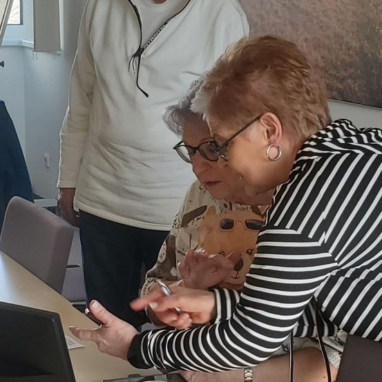 Zwei Frauen, eine mit Brille und eine andere mit Ring, untersuchen einen Laptop auf einem Tisch. Ein Mann steht hinter ihnen und trägt eine weiße Jacke und eine Brille.