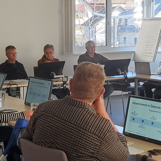 In einem Besprechungsraum sitzen mehrere Personen um einen Tisch herum, vor Bildschirmen und Monitoren. Ein Bildschirm zeigt 'eGovernment in Österreich'. Fenster im Hintergrund zeigen Gebäude.