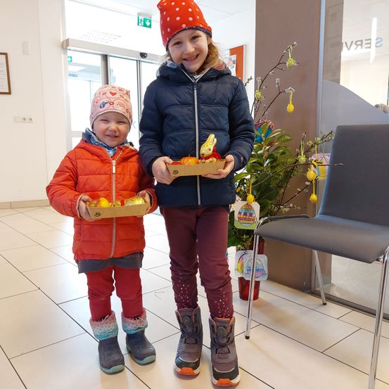 Zwei Kinder in Winterkleidung, eines hält eine Box mit Ostereiern und das andere eine Box mit Orangen. Sie stehen auf einem gefliesten Boden.