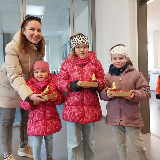 Eine Frau und drei junge Mädchen stehen in einem Raum und halten Boxen mit goldenen Gegenständen darin. Sie lächeln und posieren für ein Foto.