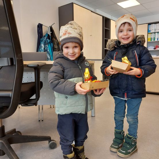 Zwei junge Jungen stehen in einem Büro, beide halten Boxen. Sie tragen Winterkleidung und lächeln in die Kamera.