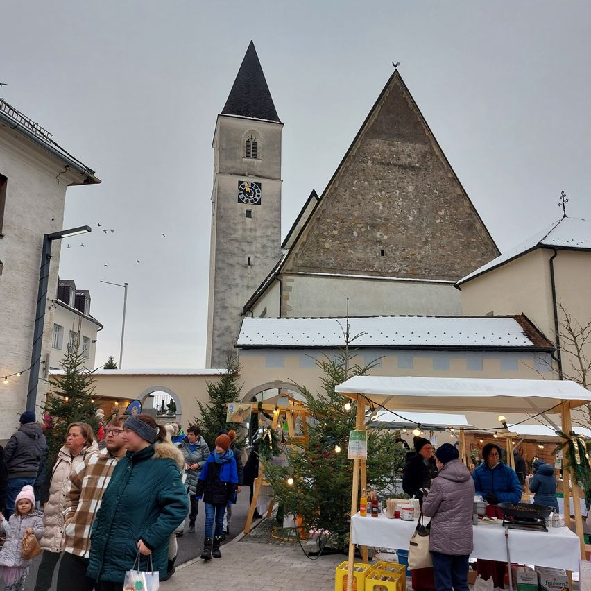Ein Weihnachtsmarkt ist vor einer Kirche mit Glockenturm aufgebaut. Leute gehen und shoppen.