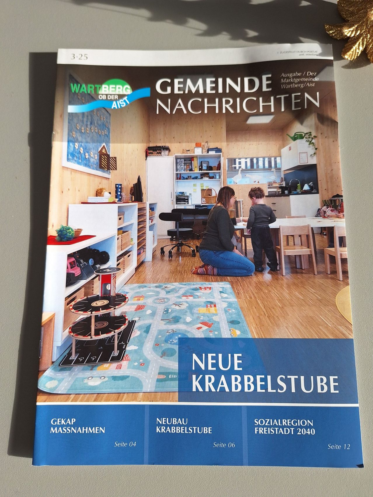 Titelseite einer Gemeindezeitung zeigt eine Frau und ein Kind in einem Raum mit Holzboden und Möbeln. Der Titel lautet Neue Krabbelstube.