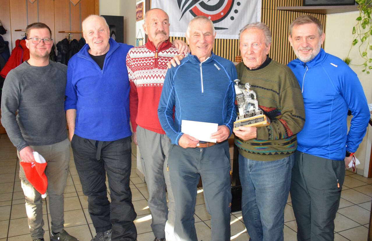 Fünf ältere Männer stehen zusammen und lächeln für ein Foto. Der Mann in der Mitte hält eine Trophäe. Hinter ihnen ist ein Banner mit einem Logo.