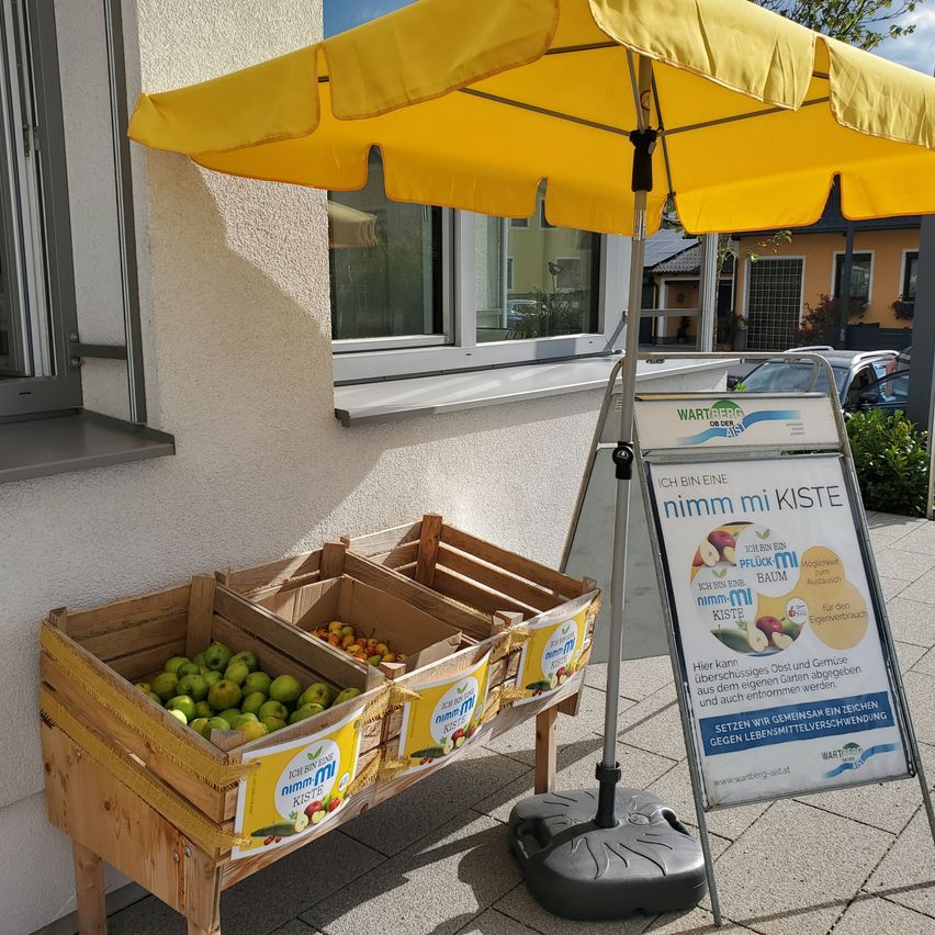 A yellow umbrella and two wooden crates filled with green apples are displayed outside a building. A sign with apples and a slogan reads, 'Take my crate.'