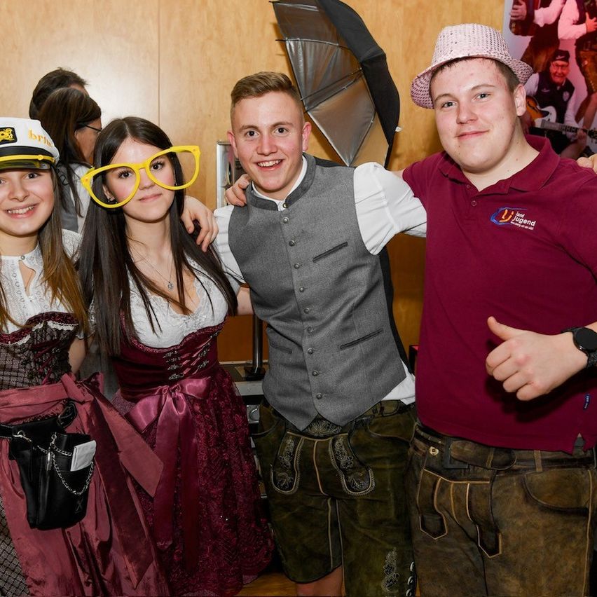 Vier junge Menschen posieren für ein Foto. Die beiden Mädchen tragen traditionelle deutsche Kleidung. Die beiden Jungen tragen Freizeitkleidung. Das Foto wurde drinnen aufgenommen.