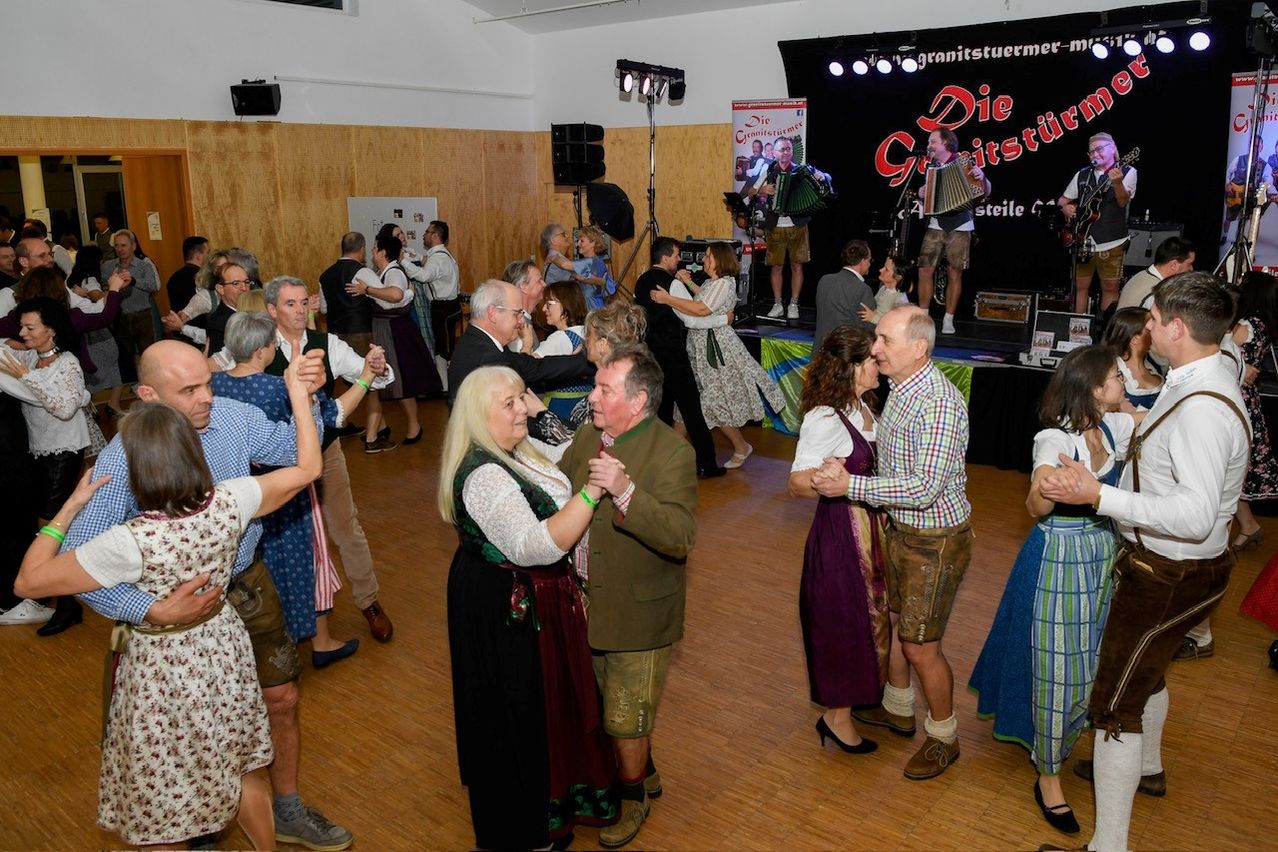 Eine Gruppe von Menschen tanzt bei einer kulturellen Veranstaltung. Ein Mann spielt Akkordeon auf der Bühne, während andere paarweise tanzen. Einige Leute stehen an der Seite und beobachten.