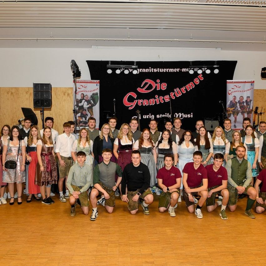 Eine Gruppe von Menschen in traditioneller Kleidung posiert für ein Foto auf einem Holzboden vor einer Bühne mit einem Banner, das 'Die Granitsturmer' liest. Sie sind von Lautsprechern und Musikinstrumenten umgeben.