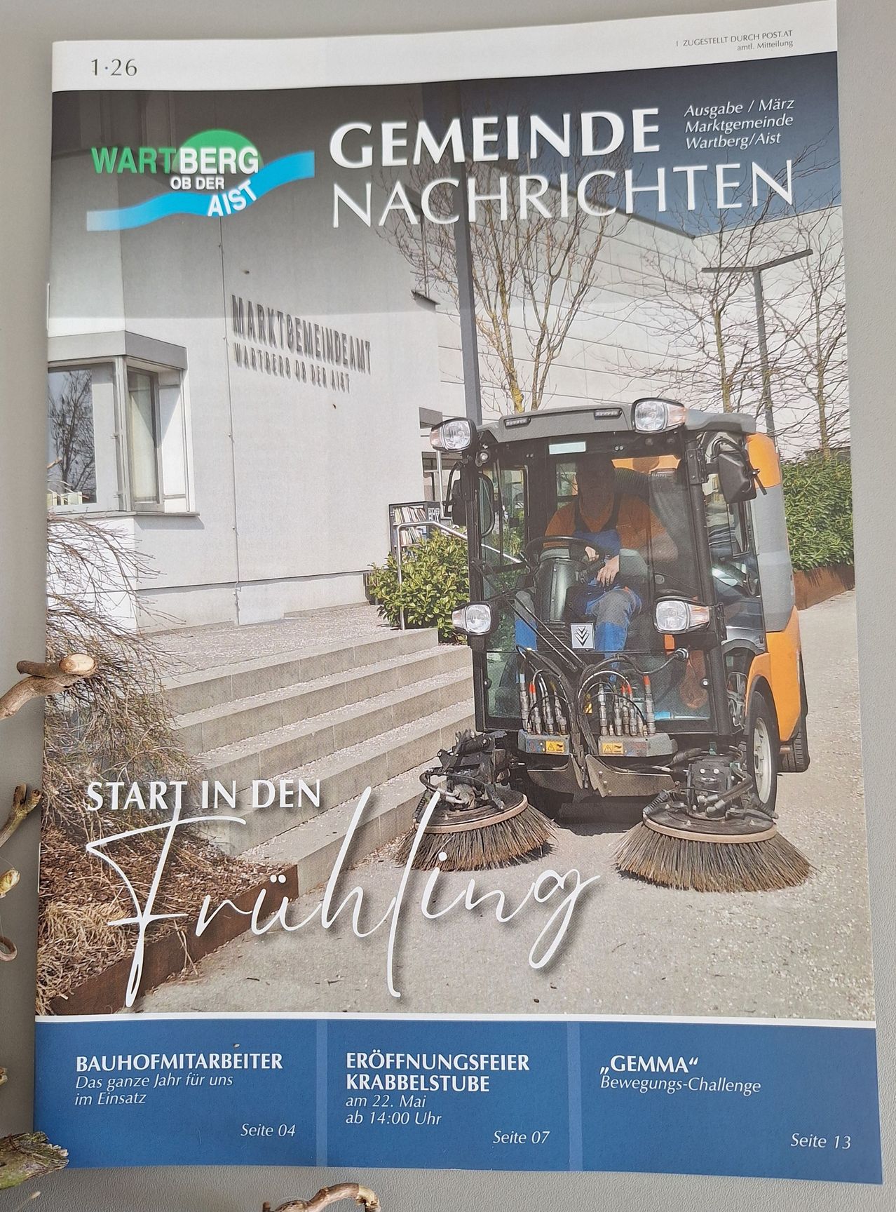 A poster featuring a person operating a street sweeper in front of a building with the text 'Start in den Frühling' and 'Wartberg Nachrichten'.