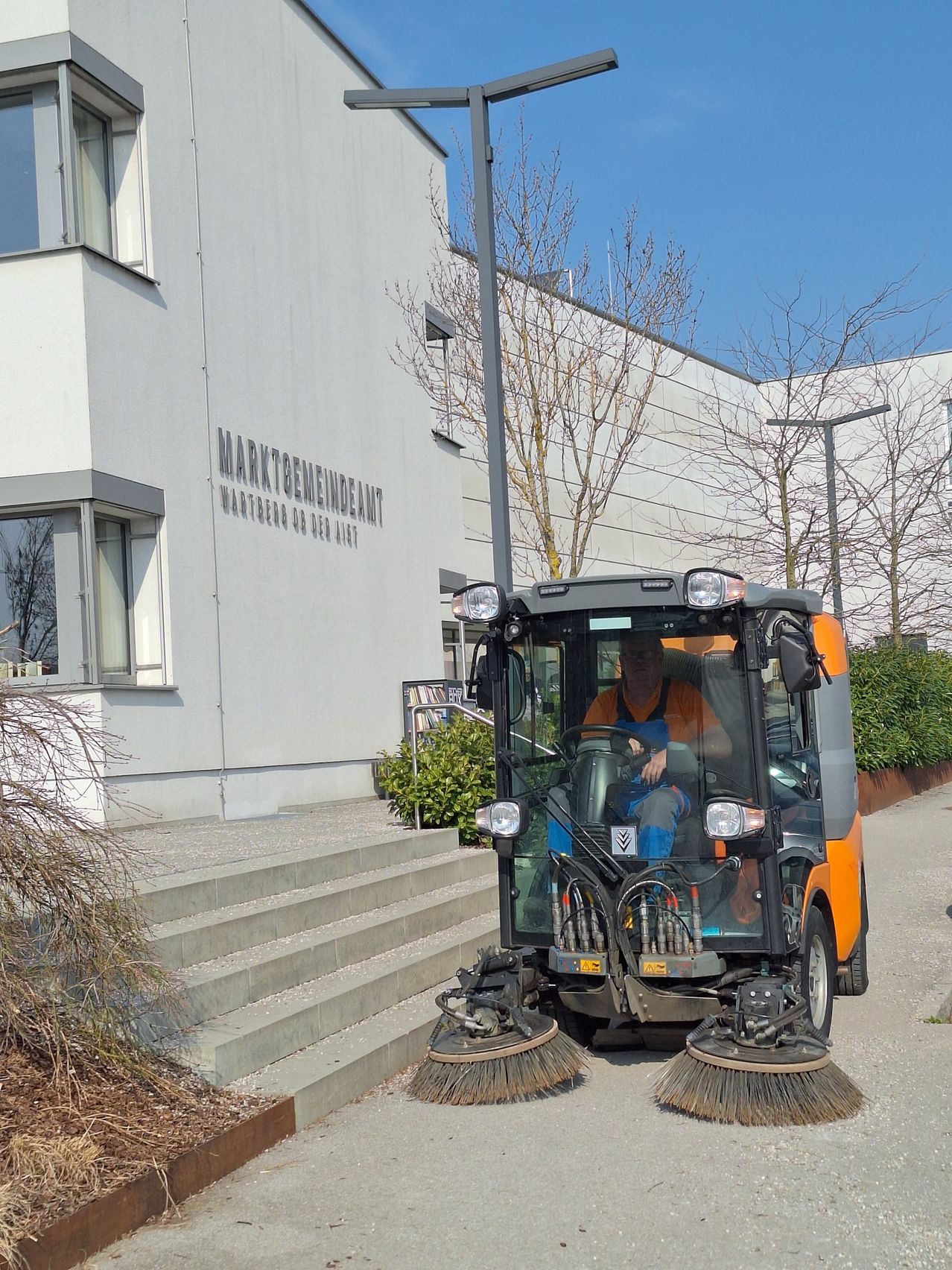 Ein Mann fährt einen Straßenkehrer vor einem weißen Gebäude mit kahlen Bäumen und Büschen.